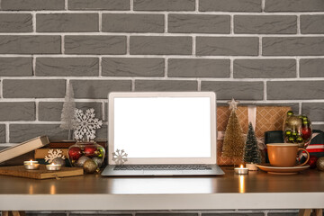 Laptop with cup, Christmas decor and presents on table near grey brick wall