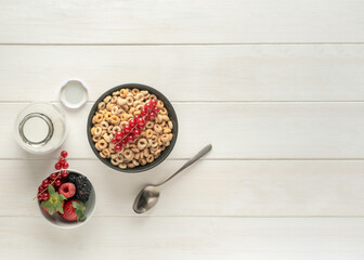 Cereal bowl with berries, spoon and milk bottle on a white table, top view, copy space.