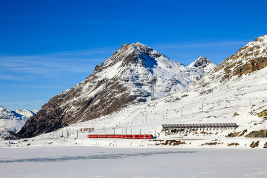 Berina, Switzerland - January 06. 2023: The red Rhaetian Rail is passing along the fronzen Lake Bianco with the Alps summit Piz Lagalb at the background