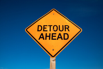 An orange diamond shaped sign stating Detour Ahead against a blue sky