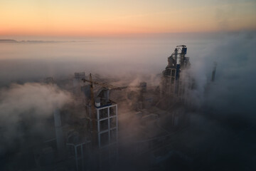 Aerial view of cement factory with high concrete plant structure and tower crane at industrial manufacturing site on foggy evening. Production and global industry concept