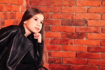 Pensive portrait brunette little girl in leather jacket posing at brick wall background, looking away. Small model thoughtful, thinking. Fashion style concept. Copy text space for advertising