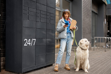 Young woman uses phone while receiving a parcel from automatic post office machine during a walk with her dog in city. Concept of modern technologies in delivery services and lifestyle © rh2010