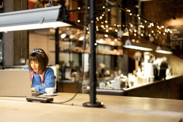 Young stylish woman works on laptop while sitting with a coffee drink at modern cafe. Concept of remote work from public place, digital freelance and modern lifestyle