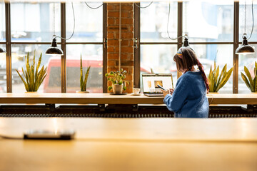 Young stylish woman works on laptop while sitting by the big window at modern coffee shop. Wide view from the backside - obrazy, fototapety, plakaty