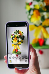 A woman's hand holds a phone ( iPhone), photographing a happiness tree with a phone, topiary with white, yellow flowers and ladybird in the center, and yellow rattan balls vertically. Copy space