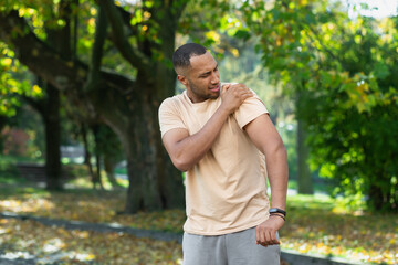 A man injured his shoulder during a fitness class, an African-American man injured himself while jogging in the park, stretches his arm and massages his sore muscles.