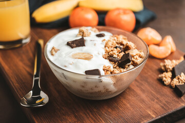 Granola cereal oatmeal with white yogurt, chocolate, banana fruit and nuts in a bowl on dark wooden board