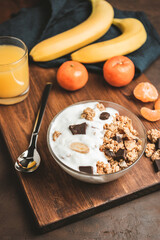 Granola cereal oatmeal with white yogurt, chocolate, banana fruit and nuts in a bowl on dark wooden board