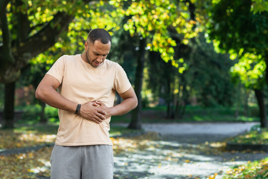 Latin American Runner Sportsman Has Severe Stomach Pain, Man Holding Hand On Side Of Stomach After Doing Exercise And Fitness In Park Outside.