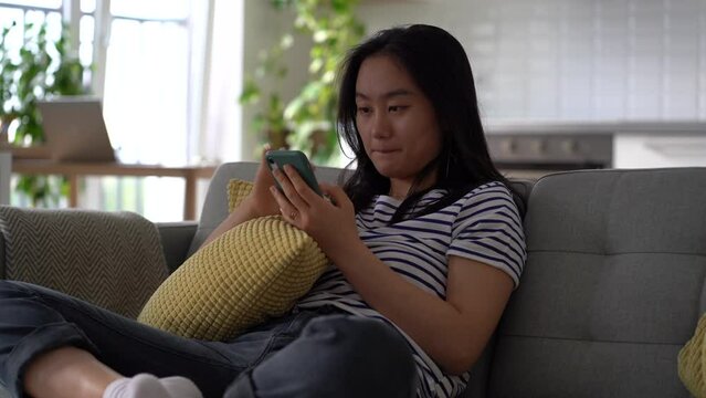 Young Asian woman holding mobile phone spying on ex on social media site while sitting on sofa at home. Korean female looking at smartphone screen paying household bills online, using banking app