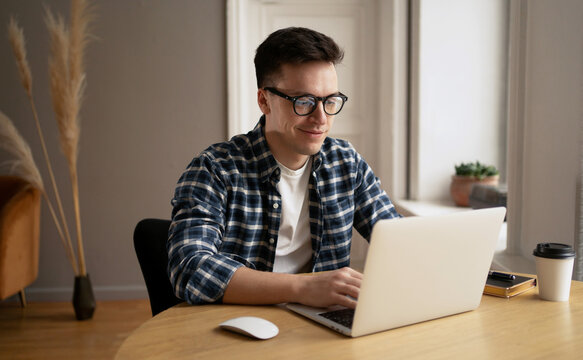 A Man In A Shirt And Glasses, A Webinar Or A Training Course Or A Computer, Studies Online From Home. Programmer Guy With Glasses Is Sitting At His Desk, Working On A Laptop, Listening To Music.