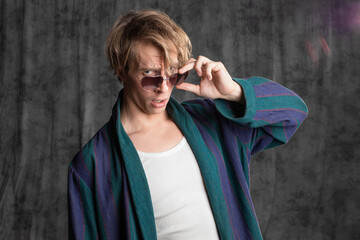 relaxed dude after a party in a housecoat with a shaggy head, posing in the studio on a gray background. Close-up with funny faces in sunglasses - obrazy, fototapety, plakaty