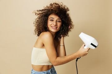 Woman dries curly afro hair with blow dryer, home beauty care styling products hair, smile on beige background - obrazy, fototapety, plakaty