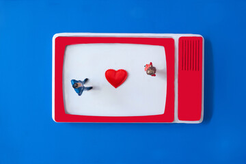 A couple standing on a white screen of a red retro tv with a red heart between them. Blue background. Minimal surreal concept for romantic movie ad. Design for Valentine or wedding card or banner. Top