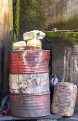 Full view of old and dented red and white oil barrel with empty plastic containers piled on top