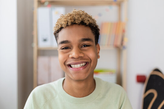 Portrait Of Smiling Young Latin American Teenage Boy Looking At Camera Indoors. POV Screen View Of Millennial Man On Laptop Video Call In His Room