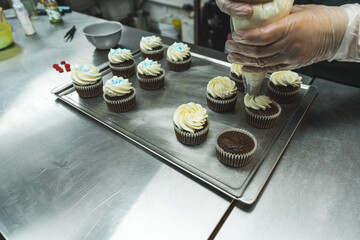 Closeup shot of person adding vanilla buttercream frosting with pastry bag onto chocolate muffins placed on stainless steel baking tray. High quality photo