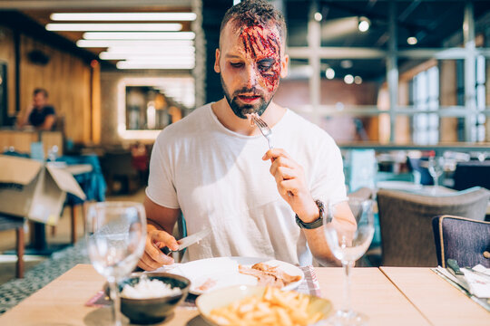 Man with makeup on his face with wounds and blood. Eating at the table