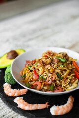 Wok, noodles, udon with shrimp and vegetables, on black plate and a wooden white background