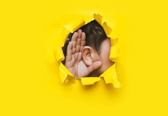 Close-up of a man's ear and left hand through a torn hole in the paper. Yellow background, copy...