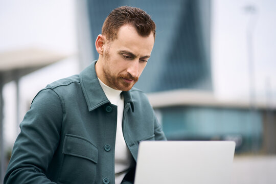 Pensive handsome programmer using laptop computer working freelance project sitting at workplace. Young businessman watching training courses, planning startup outdoors 