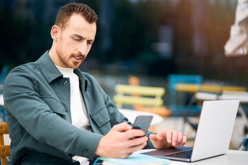 Young confident businessman using laptop holding mobile phone working online outdoors. Portrait of handsome successful freelancer checking email sitting at workplace. Technology concept 