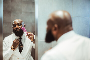 A capture of a mature well dressed fully bearded bald black male in a white vest with a polka-dot necktie combing his beard with a pink comb in front of a narrow mirror, in a grey-stoned restroom