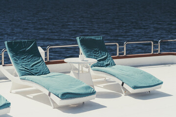 A wide-angle shot of two lounge chairs with blue cushions on top of them and a small white color coffee table on the upper deck of a luxurious sailing yacht on the sea, in the Maldives island