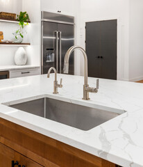 Detail image of faucet, sink, and island in new kitchen. Background features stainless steel refrigerator and modern shelving.