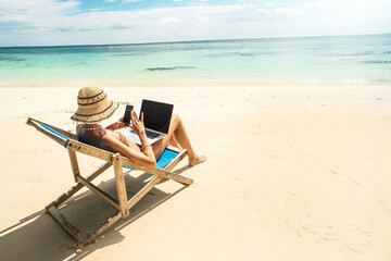 Photo fo a woman in summer hat using mobile phone and laptop computer, working while sitting on a beautiful tropical beach. Digital nomad. - 559255949