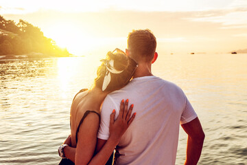 Back view of romantic couple looking at the golden sunset, relaxing at the beach, enjoying tropical honeymoon. Copy space. Tourism.