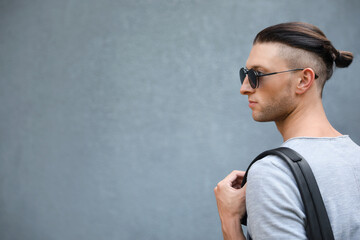 Handsome young man in stylish sunglasses with backpack near grey wall, space for text