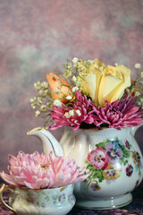 A floral teapot and teacup with an arrangement of fresh roses, mums, and baby's breath flowers in pink, yellow, and white
