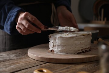 Woman smearing sponge cake with cream at wooden table, closeup