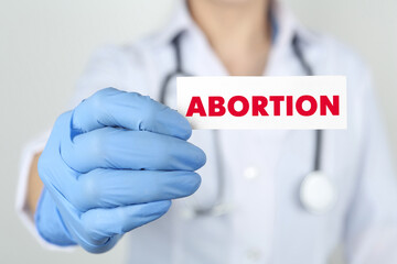 Doctor holding paper card with word Abortion on light grey background, closeup