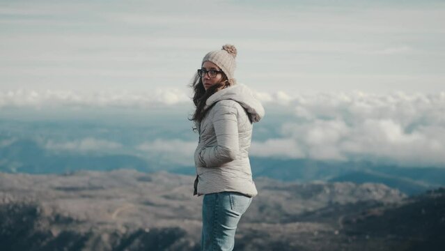 Young thoughtful woman in a gray jacket, hat, and scarf contemplates a stunning mountain landscape on a hike. Slow motion, medium shot. Ideal for themes of nature, outdoor adventure, and contemplation