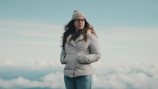 A beautiful young woman in a gray jacket and glasses contemplates the beauty of nature while taking in the stunning panoramic views of the mountains and clouds. Medium shot filmed in slow motion.