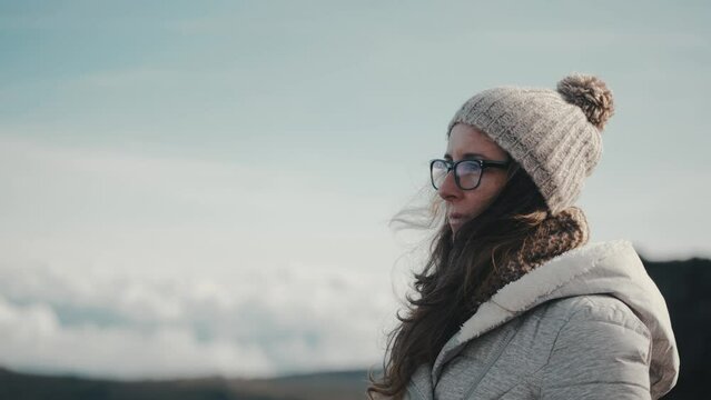 Slow motion close-up of a thoughtful inspired woman enjoying the beauty of nature in the mountains. Young, millennial, and wearing glasses, a gray jacket, hat, and scarf. Travel and adventure visuals.