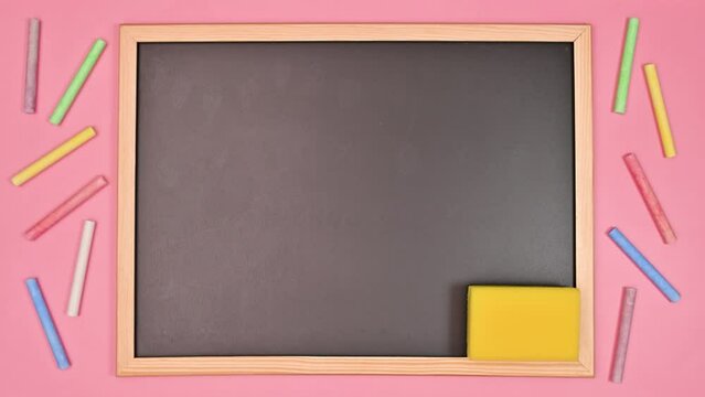 School blackboard with sponge and chalks appear on pastel pink background. Flat lay stop motion