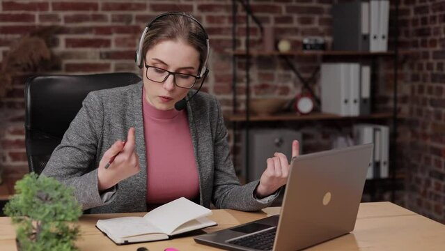 Female young brunette woman in glasses and modern headset with microphone holding online meeting conversation and using laptop writing notes in office, studying distantly, discussing project.