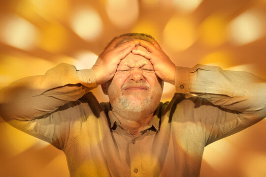 An elderly man is holding his head with both hands, he is suffering. Bright light surrounds him. 