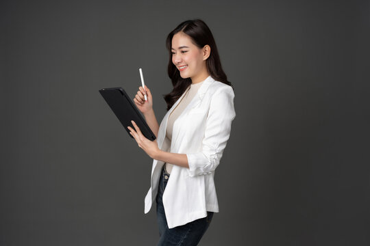 A female executive wears a long-sleeved white coat. Taking pictures against a gray background in the studio She stood at work. He looked at the tablet in his hand and held the pen with a smile.