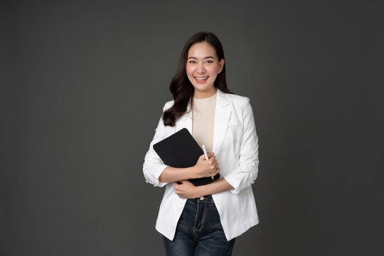 Portrait Of Long Haired Female Executive With Gray Background In Studio Wear A Long-sleeved White Shirt. Holding A Notebook Tablet And A Pen, Smiling Beautifully.