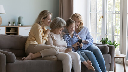 Happy little daughter kid, young mom, middle aged grandma, elderly great grandmother using mobile phone for family video call, sitting on sofa together, looking at cellphone screen, smiling