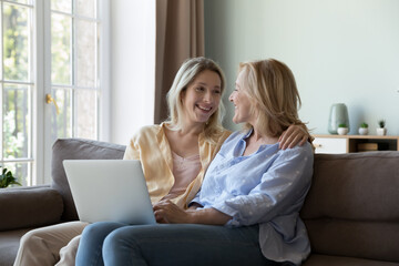 Happy cute senior mom and adult daughter chatting at laptop, sitting on home sofa, holding digital gadget on lap, talking, laughing, enjoying leisure, Internet online communication