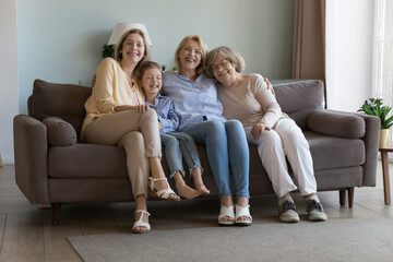 Cheerful cute girls and women of four family generations sitting together on home couch, laughing, hugging with love, affection, looking at camera. Kid, mother, grandma, great grandmother portrait