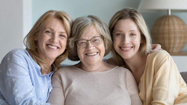Cheerful pretty women of different ages, generations sitting close together, looking at camera, smiling, enjoying family relationships, meeting at home, leisure, mothers day