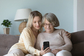 Positive adult granddaughter woman teaching old grandmother to use online app on mobile phone,...