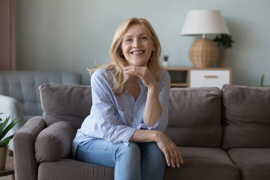 Happy blond middle aged attractive woman sitting on comfortable couch at home, touching face, leaning chin on hand, looking at camera with toothy smile, laughing
